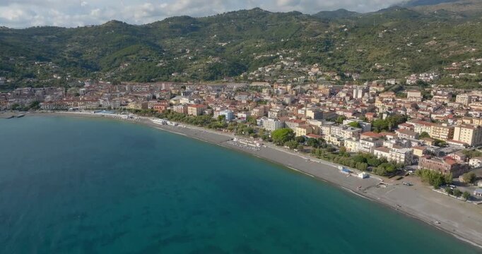 Panorama of a vibrant coastal town curving along a deep blue bay. Dense buildings sit between a long beach and green hills. Aerial view of seafront of Sapri, in province of Salerno, Campania, Italy.