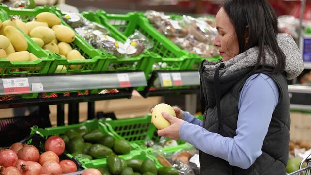 A vertical video of a middle-aged Caucasian woman selecting mangoes at a local supermarket and placing them in her shopping basket.
