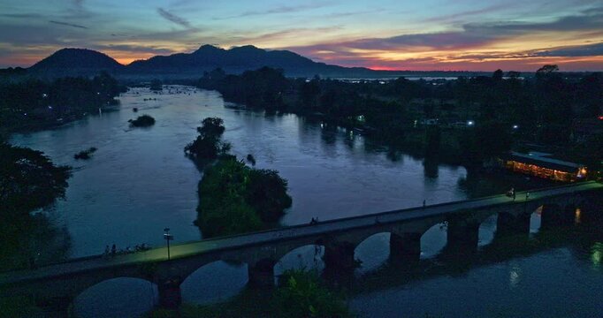 Drone view of the iconic red bridge connecting a small island at Li Phi Waterfall on the Mekong River. The warm sunset light reflects across the river valley, highlighting the harmony between natura