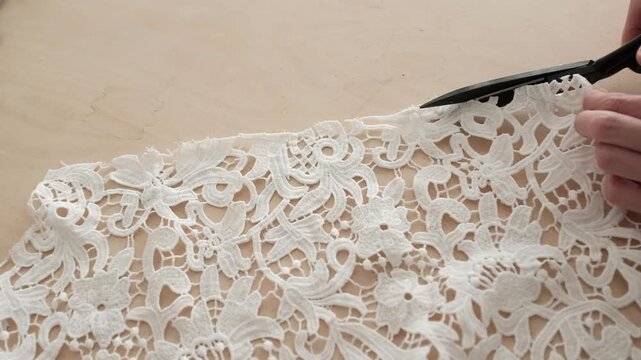Close up of female hands carefully cuts white lace fabric with scissors. Minimal sewing environment, highlighting manual sewing manufacture. Working process in a small business sewing workshop.