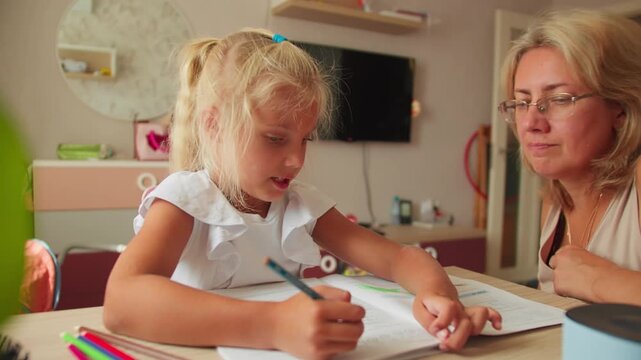 Child writing in a workbook while mother assists nearby. Home education and parent-child interaction