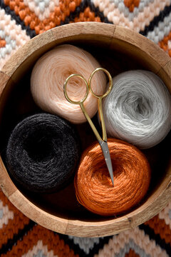 Top view of yarn balls in wooden bowl and vintage scissors on bargello needlepoint blanket, colorful wool thread for embroidery, cozy handmade geometric flame stitch craft background