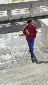 Vertical video: Walking red hoodie dodging puddle, going up steps at plaza, beanie holding coffee