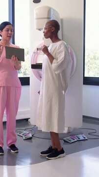 Vertical video: Tech tapping tablet in pink scrubs guiding patient in white gown at mammogram unit