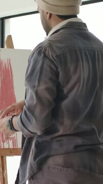 Vertical video: Reaching for brush, Indian artist in beanie applying red marks at easel to refine