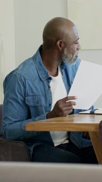 Vertical video: Reading paper, man in denim turning to sound and smiling in dining nook, glasses