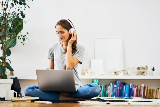 Relaxed young woman at home sitting on the floor using laptop and listening to music