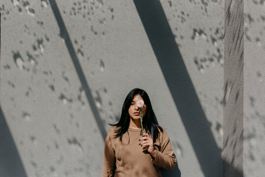 Woman holding flower in front of eye while standing against concrete wall on sunny day