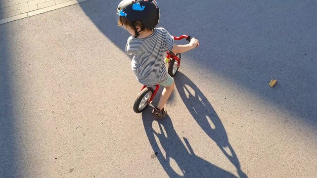 High-angle view of a child wearing a safety helmet while learning to ride a red balance bike on an asphalt path, showcasing childhood skills and outdoor fun at sunset