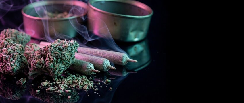 Close-up of cannabis buds, ground marijuana, and pre-rolled joints with smoke and colorful lighting on a dark reflective surface.