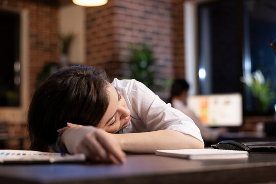 Tired businesswoman takes powernap at her workstation, resting briefly amid her demanding schedule in late night office setting. Caucasian entrepreneur dozes off at desk, exhausted from her workload.