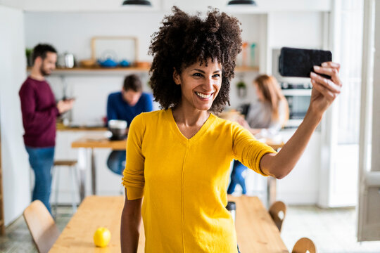 Happy woman taking a selfie at home with friends in background