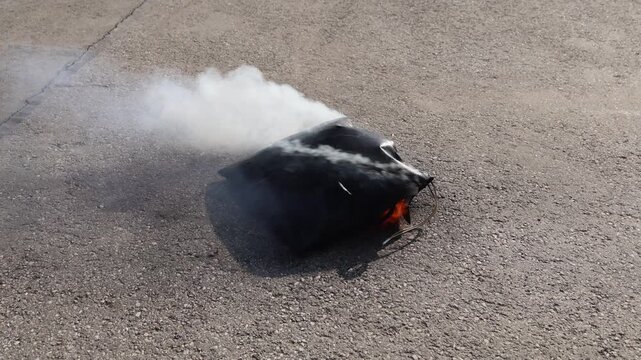 Heat-resistant safety bag containing a lithium battery failure with visible flames and smoke escaping, demonstrating fire containment on an asphalt surface