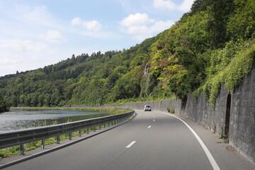 Scenic road curves alongside a river and lush green forested hillside with a single vehicle