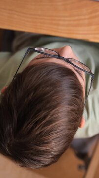 Vertical video young person wearing modern eyeglasses focusing intently while working or studying at a wooden desk, seen from a high angle view emphasizing concentration and dedication