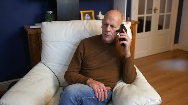 A mature senior bald man listening and talking on his phone. The mature male is stiing at home wearing jeans using his cellphone