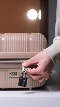 Vertical video person's hand attaching a black combination padlock to zipper pulls on a beige hardshell suitcase, ensuring security for valuables during travel and transport