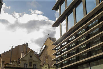 Historic rooflines meeting modern glass corner under dramatic sky, textured masonry and gabled tiles contrast with sleek © Alena