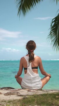 Woman in white dress meditate outdoor on fresh air. Female relax sitting in lotus position on sea coast under coconut tree leaf in sunny day. Self care, mental relaxation exercises, feels no stress