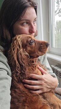 A girl with a dog in her arms looks out the window