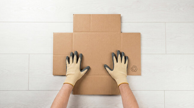 Gloved Hands Flattening Cardboard Box with Recycling Symbol for Recycling on White Floor &mdash; Top View
