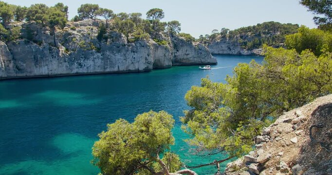 Scenic view of Calanque de Port-Miou framed by pine trees, with motorboat crossing deep blue and turquoise Mediterranean water along rocky cliffs
