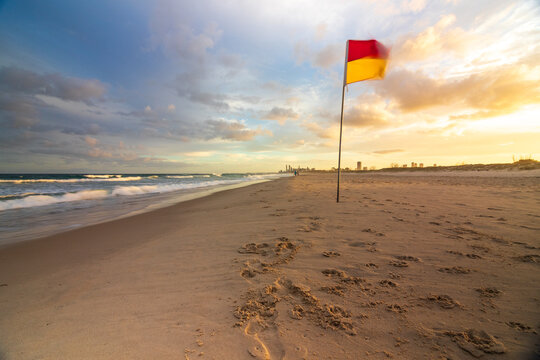 Red and yellow lifeguard flag blowing in wind on sandy beach with tide out and sunset sky