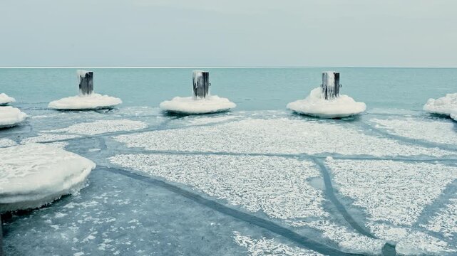 Frozen lake shows round patches of ice and wooden posts during winter. The icy surface reflects the light from the sky above with no signs of movement or activity.