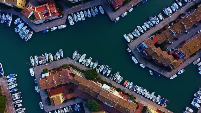 Multiple boats are anchored in a marina with narrow water channels. Buildings line the sides. This scene is set in a coastal area under clear skies during daylight.