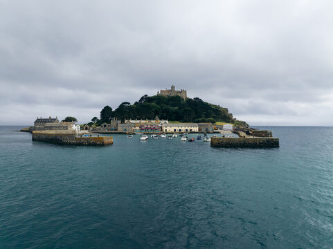 Aerial view of St Michael's Mount rising majestically from the turquoise sea under a blanket of grey sky, its ancient stone walls steeped in history, Marazion, England, United Kingdom.