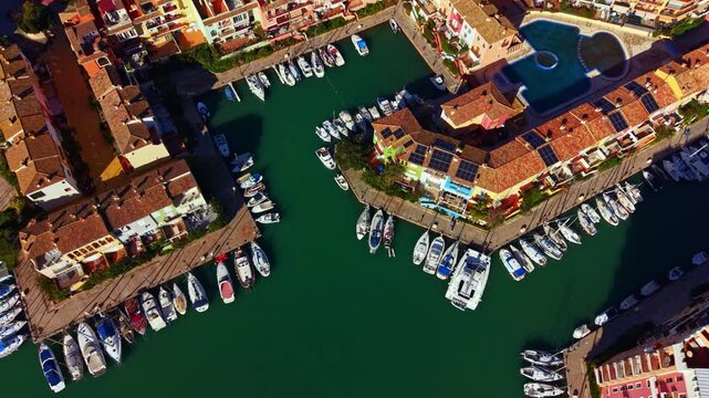 View from above shows a marina filled with boats. Buildings line the water's edge and a pool is visible among the structures. The scene is bright and the water is clear.