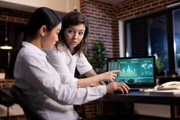 Diverse women collaborate and discuss strategy ideas in a late night office meeting, using financial graphs on laptop. The scene portrays a professional and focused atmosphere with dim lighting. © DC Studio