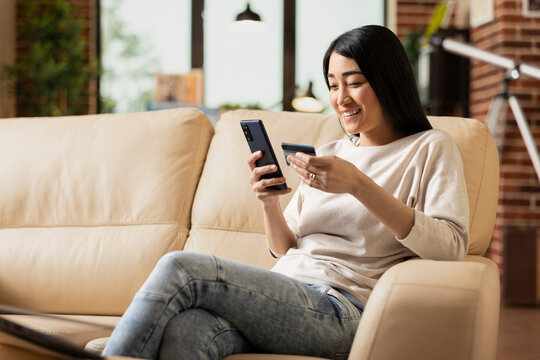 Remote asian manager happily using cellphone and debit card for mobile banking. Confident businesswoman seated on sofa in home office, accessing online tools to complete financial tasks.