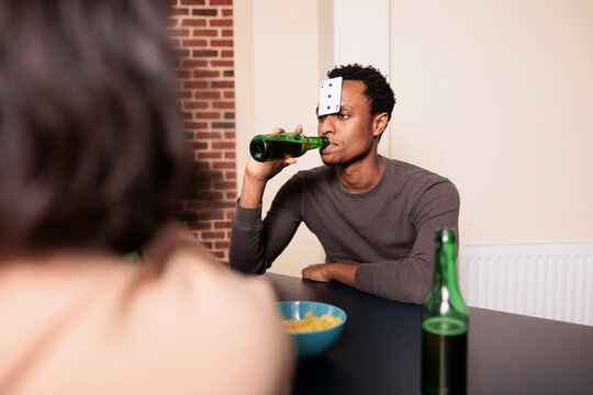 Confident young adult man with plastic card on forehead drinking beer while playing charades with friends. Person sitting at table in living room while enjoying fun leisure activity.