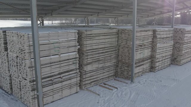 Large stacks of flat panels or boards stored under a metal-roof shelter in a snowy outdoor yard, showing industrial organization and winter conditions