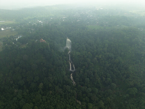 Aerial view of cascading waterfalls plunge through the dense, verdant forest canopy, a misty veil hanging in the air, Curugsewu, Kendal Regency, Central Java, Indonesia.