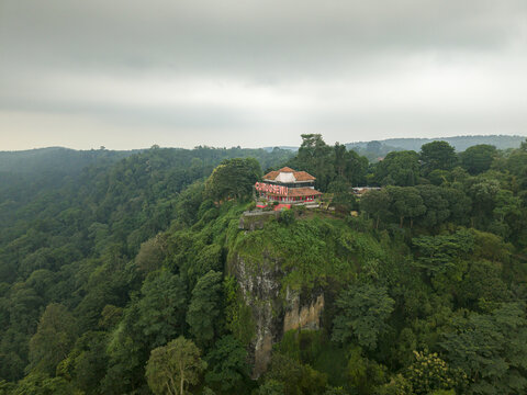 Aerial view of a vibrant red-roofed building perched atop a dramatic cliff surrounded by dense, lush green forest under a cloudy sky, Curugsewu, Central Java, Indonesia.