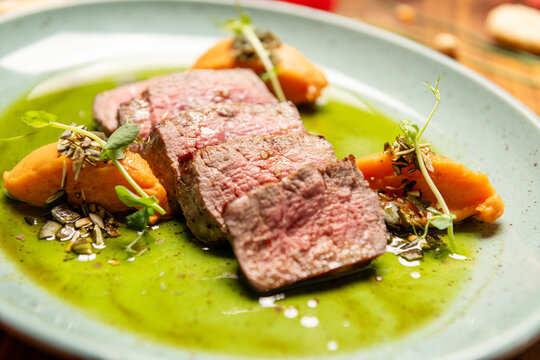 Juicy beef steak slices on rich green sauce with carrot puree and microgreens, beautifully plated on ceramic plate, rustic wooden table