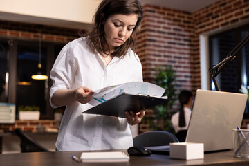Fototapeta premium Professional caucasian woman stands at her desk, holding a clipboard and flipping through documents. She carefully reviews company research data in a modern brick walled office late at night.