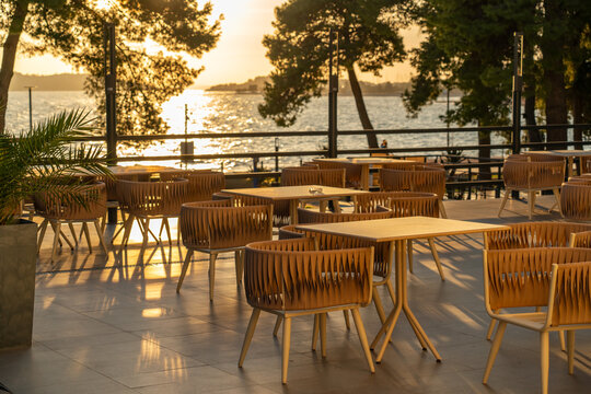 Outdoor seaside restaurant terrace with empty tables and chairs during golden hour, overlooking the water. The image represents hospitality, lifestyle, and travel dining experiences