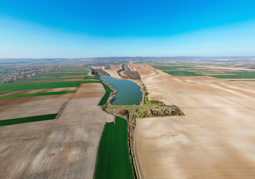 Aerial view of a serene lake nestled amidst the contrasting textures of cultivated fields, creating a patchwork of earthy browns and vibrant greens, Mandelos, Vojvodina, Serbia.