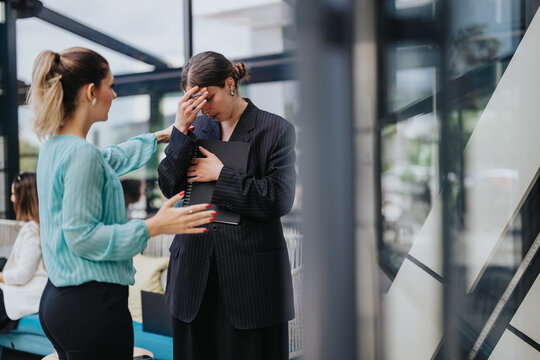 A distressed businesswoman is comforted by a female coworker while holding a folder. The office scene shows workplace collaboration and emotional support between colleagues.