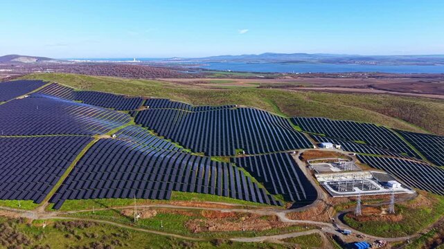 Rows of solar panels create a vast field on a hillside overlooking a lake with mountains in the background on a clear day.