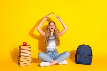 Young woman sitting with book and a backpack on a yellow background showcasing a cheerful pose