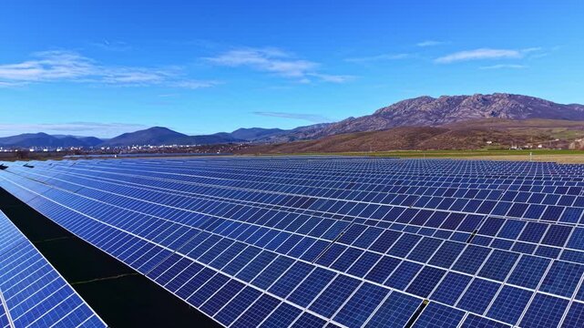 Large solar panel array spreads across land with mountains in the background. The scene shows clear blue sky and the vastness of the energy project during daylight.