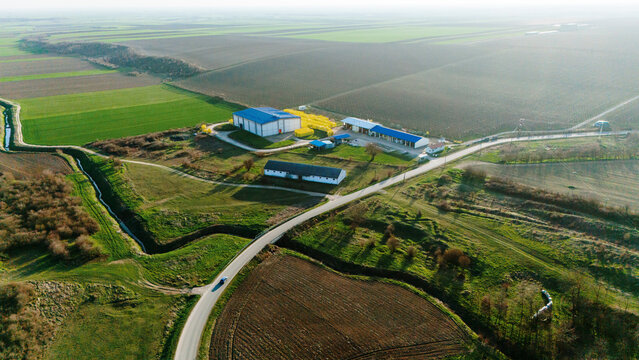 Aerial view of the vibrant patchwork of agricultural fields meeting modern industrial buildings under a soft, hazy sky, Jazak, Vojvodina, Serbia.