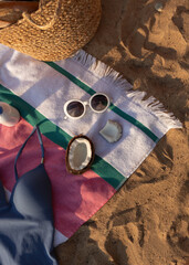 Obraz premium Jute beach bag with straw hat, swimsuit, coconut and sunglasses on the striped towel on the sand. Summer vacation lifestyle concept
