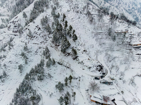 Aerial view of snow-laden trees and winding roads create a stark, monochrome landscape, punctuated by the dark silhouettes of buildings, Pir Chanasi, Azad Kashmir, Pakistan.