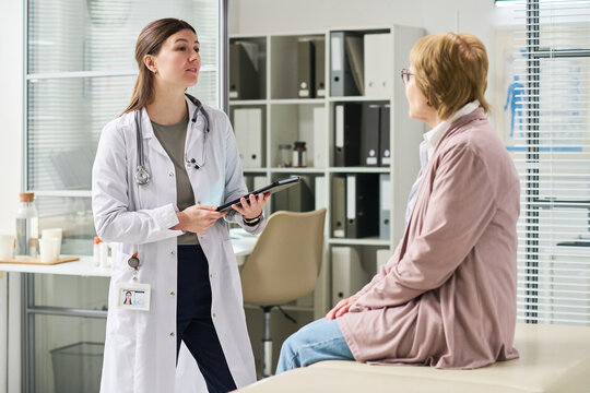 Young female doctor holding digital tablet speaking with senior woman sitting on examination table in medical office during consultation