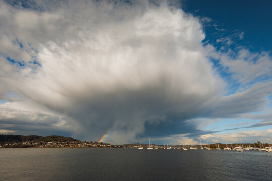 Large storm cloud formation with vibrant rainbow over lake with distant town and moored sailboats